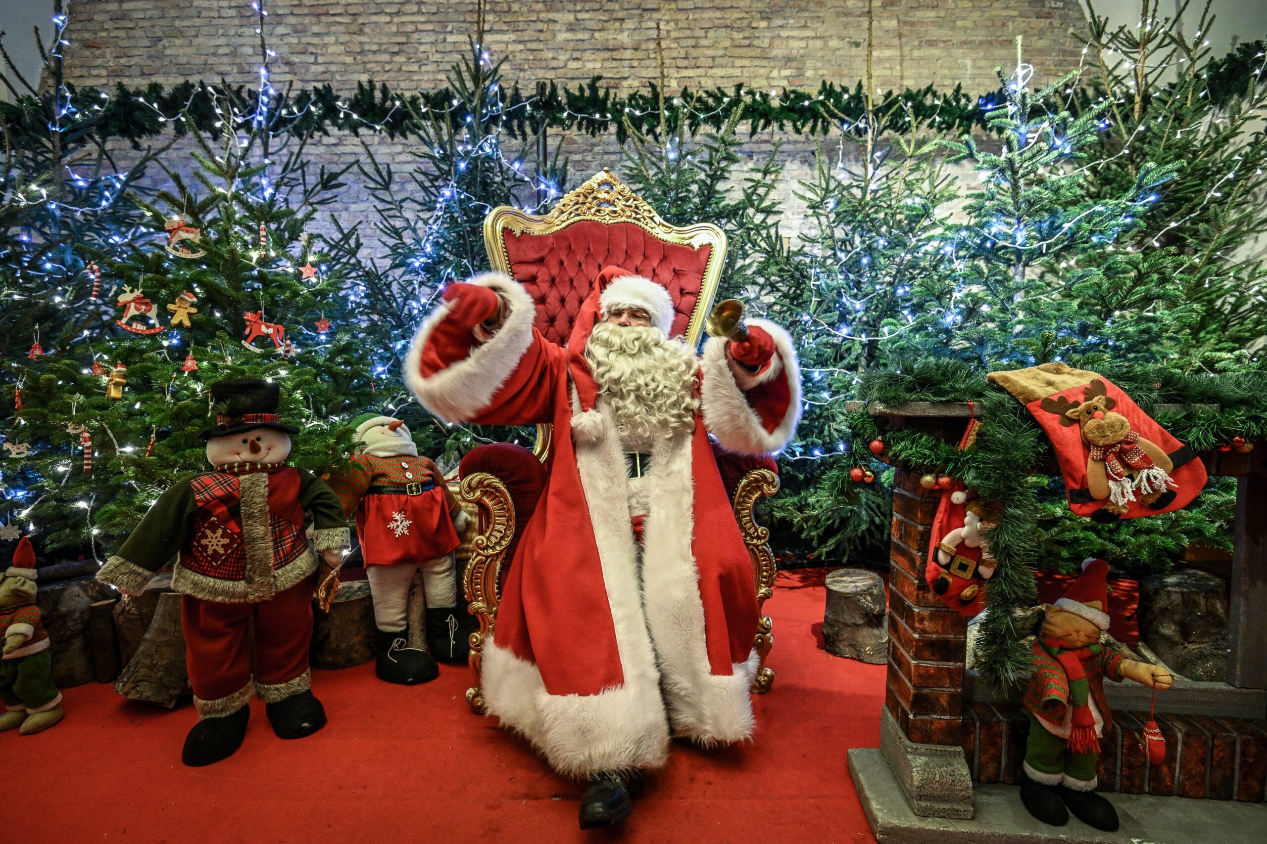 Święty Mikołaj, siedząc na ozdobnym tronie, daje dziecku prezent w postaci lizaka. W tle zdjęcia znajduje się choinka i świąteczne lampki, a na dalszym planie Gdańskie budynki.